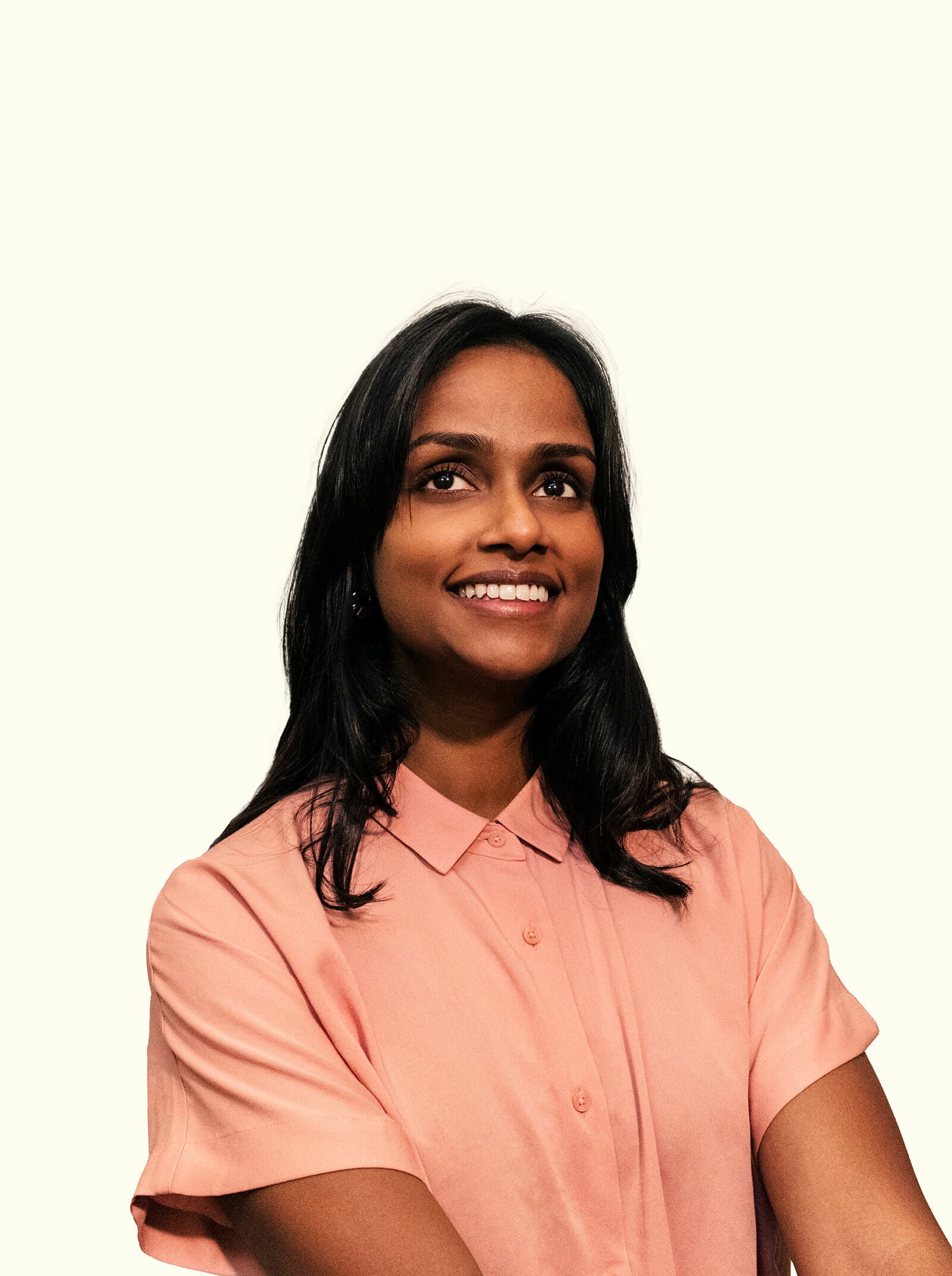 A person with long black hair, wearing a light pink button-up shirt, is smiling while looking slightly upwards. The background is a plain, light-colored backdrop.