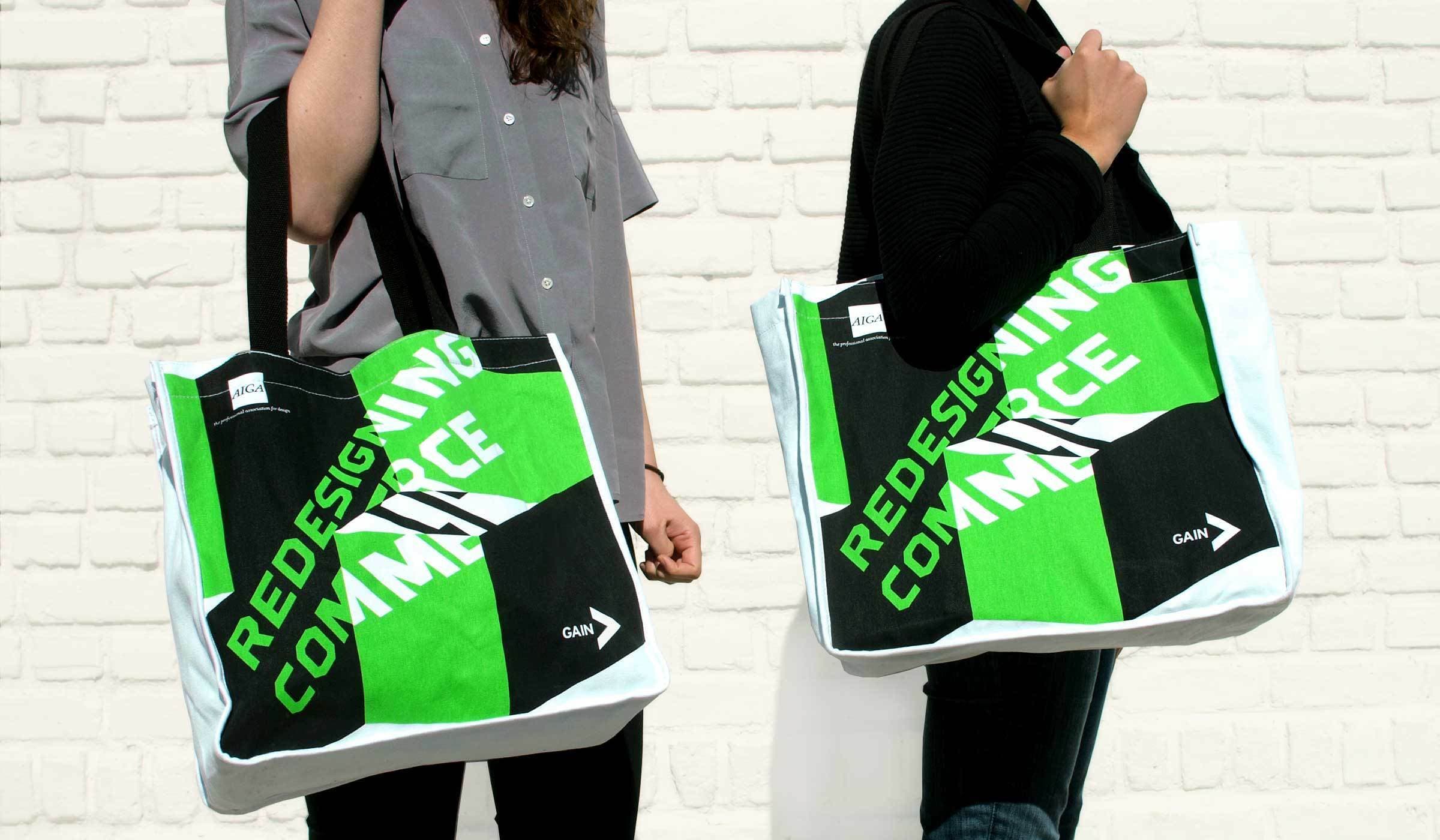 Two individuals are standing against a white brick wall, each holding a large tote bag. The tote bags are white with a bold green and black design stating "REDESIGNING COMMERCE" in bright green letters and a smaller "GAIN" logo in white on the bottom right.