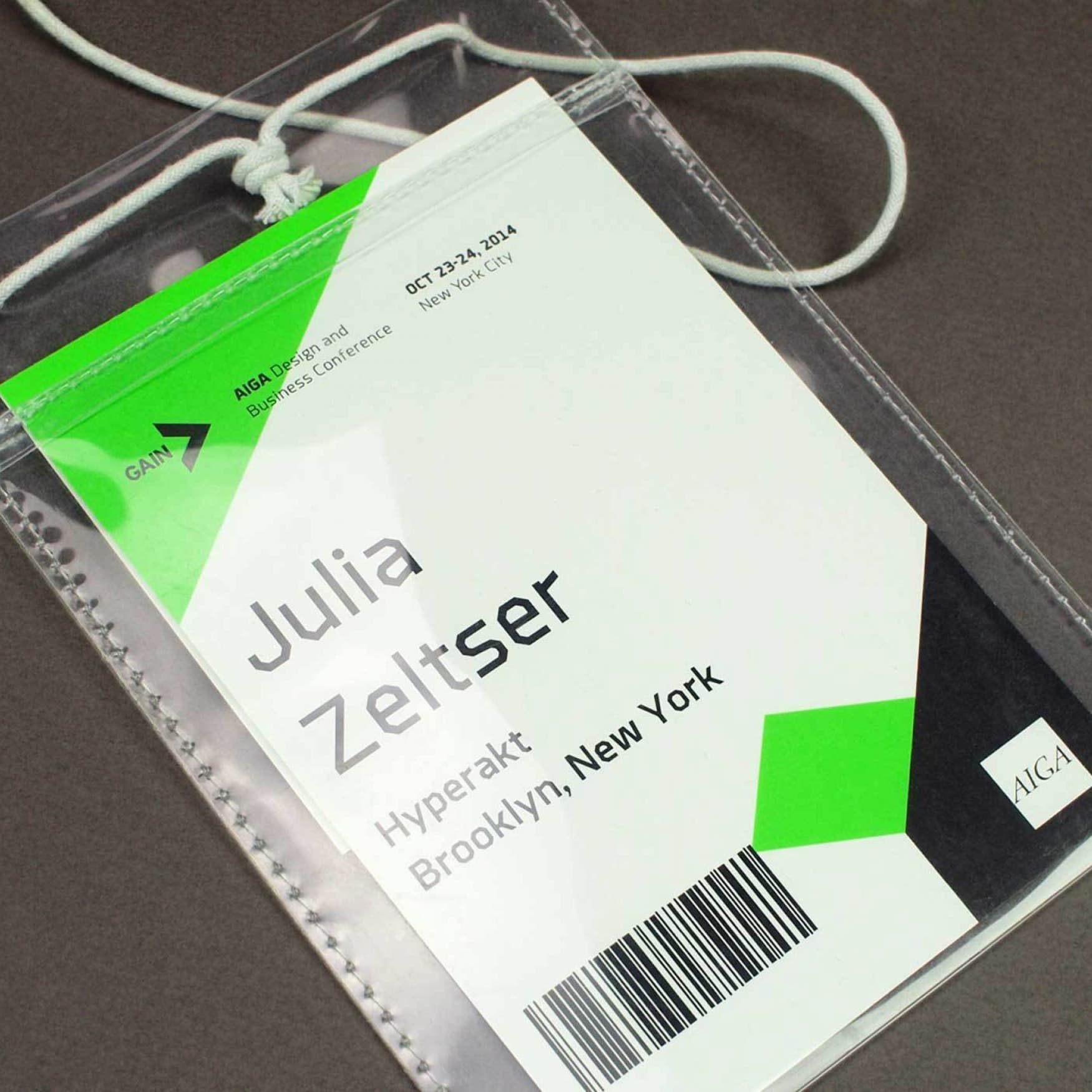 A name tag lays on a dark surface. It reads "GAIN: Design for Social Value" and "AIGA Business Conference" at the top left, "Julia Zeltser" in the center, and "Hyperakt Brooklyn, New York" at the bottom. It includes a barcode, date, and conference location.