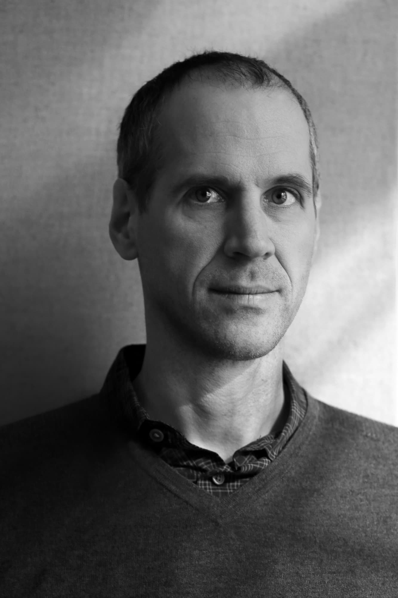 Black and white portrait of a middle-aged man with short hair, light stubble, and a focused expression. He is facing the camera, wearing a collared shirt and a sweater. The background is plain and softly lit, creating a calm and introspective atmosphere.