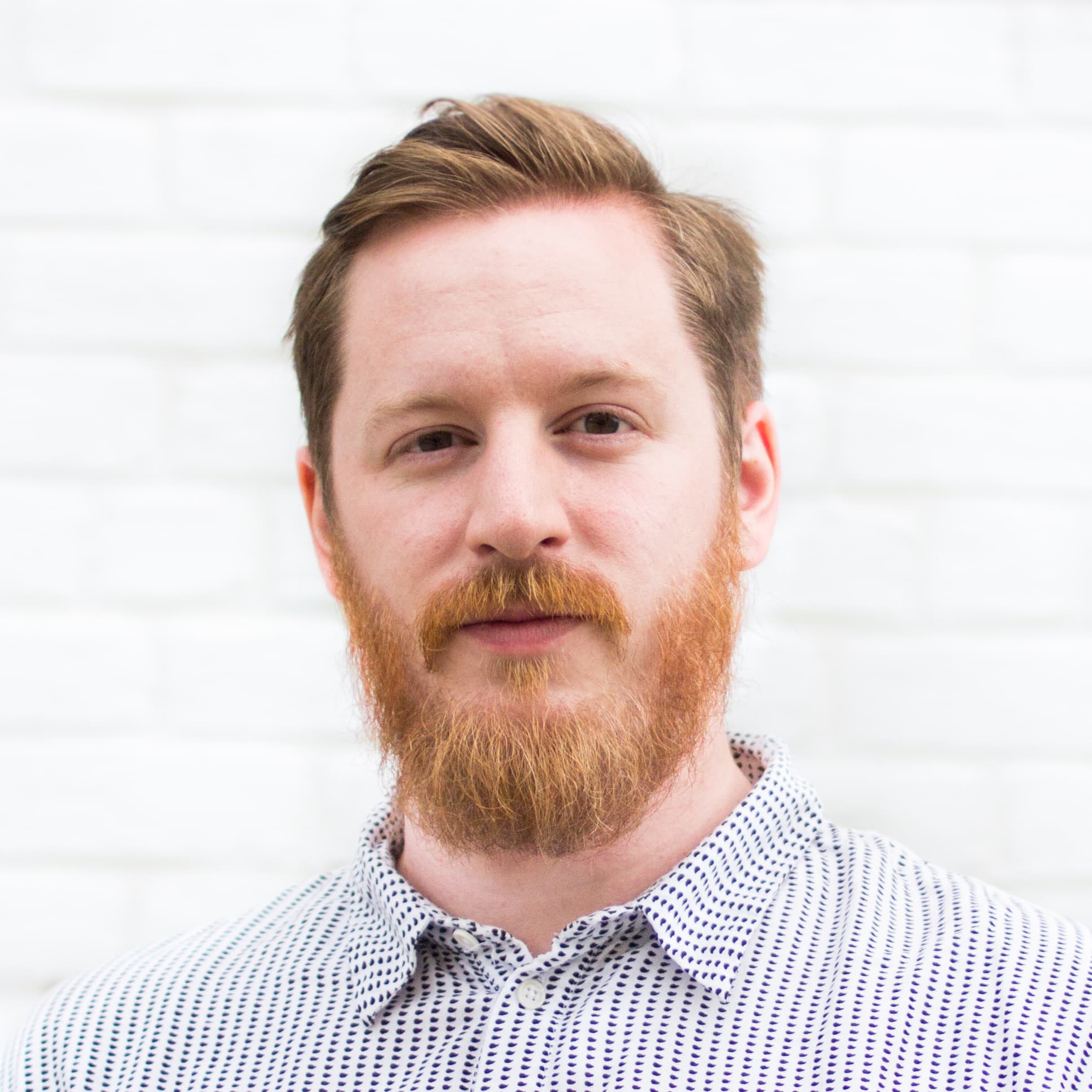 A man with light skin, ginger hair, and a full beard stands against a white brick wall. He is wearing a white, patterned button-up shirt and has a slight smile.