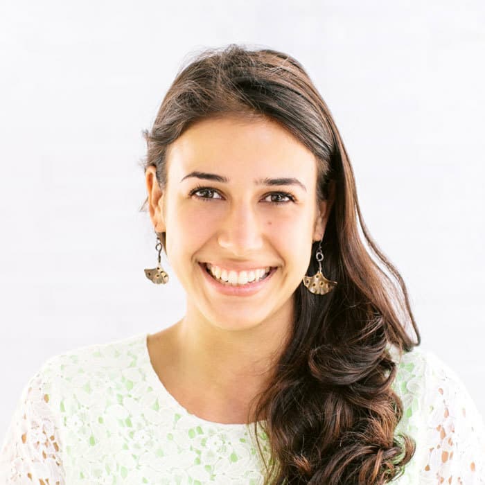 A woman with long, wavy brown hair smiles broadly at the camera. She is wearing a white floral lace top and dangly earrings shaped like cats. The background is bright and slightly blurred, emphasizing her cheerful expression.