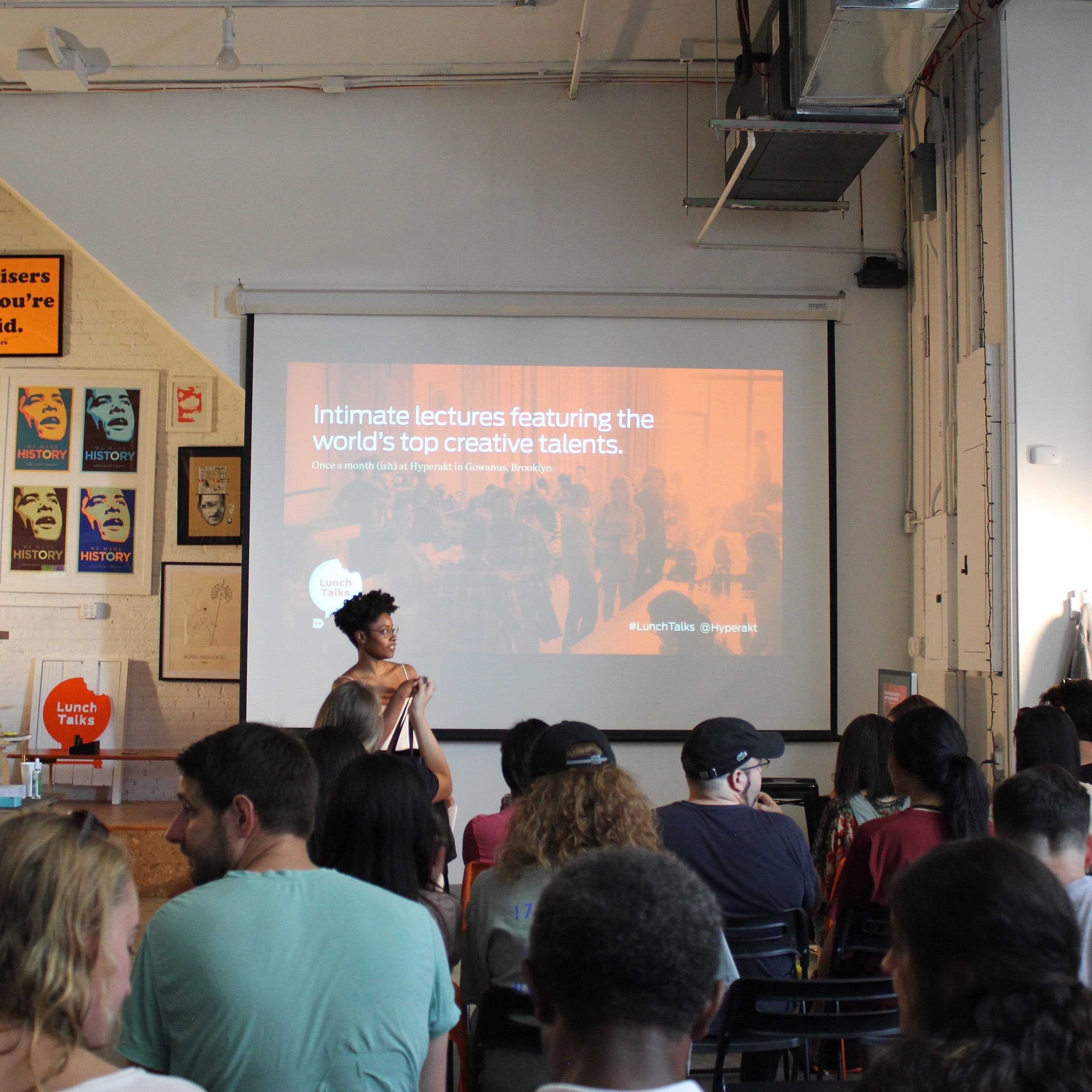 A room filled with people seated and facing a screen displaying a presentation titled "Intimate lectures featuring the world's top creative talents." The room is decorated with various posters and art. One person at the front is gesturing while speaking.