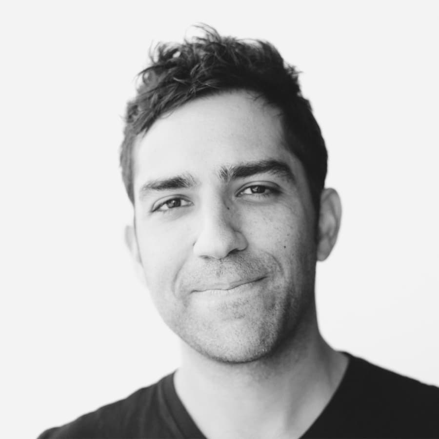 Black and white portrait of a person smiling with short, tousled hair and wearing a dark-colored, V-neck shirt. The background is uniformly white, highlighting their facial features.