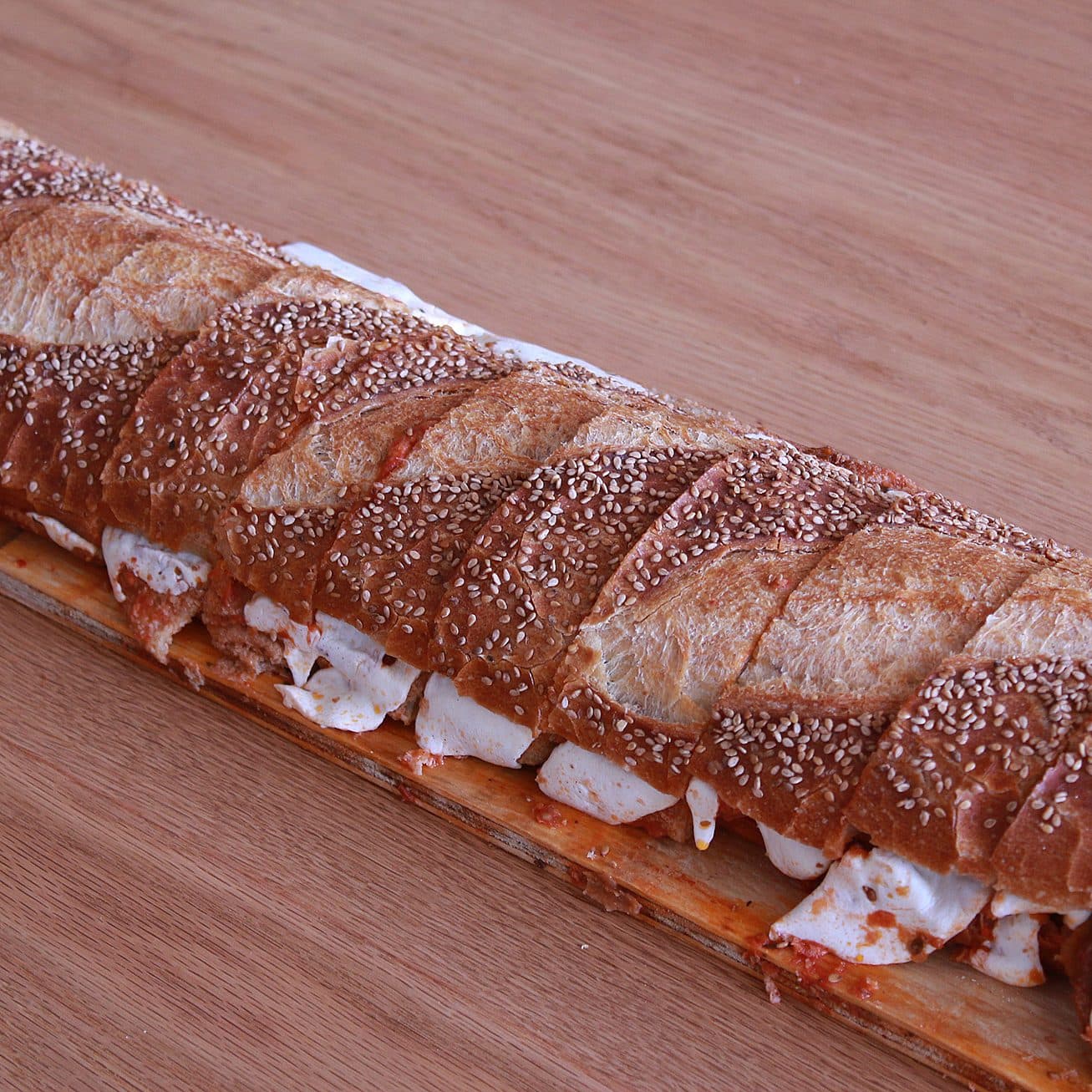 A long, crusty baguette sandwich filled with cheese and a reddish spread, possibly tomato-based. The bread is sprinkled with sesame seeds and placed on a wooden surface. The sandwich is sliced into portions.