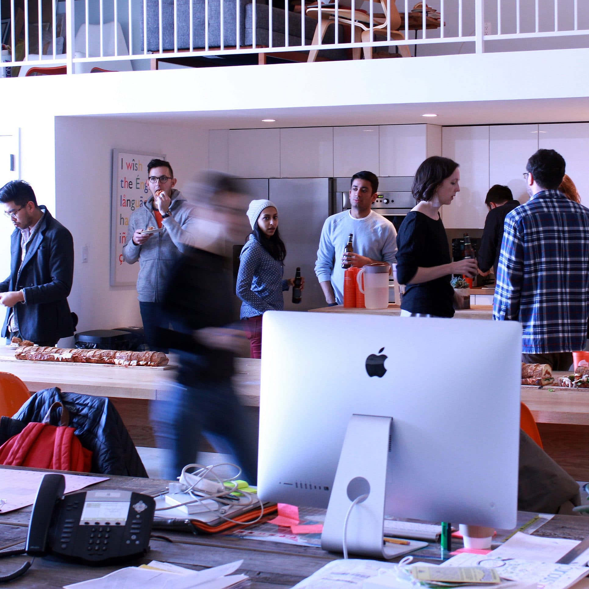 A group of people are gathered in a modern, open office kitchen area, socializing and eating. The room has white walls, a long wooden table, an office desk with a computer and a telephone in the foreground, and an upstairs loft visible in the background.