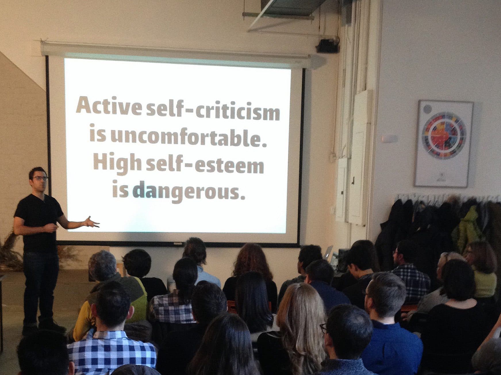 A person stands in front of an audience in a classroom, gesturing towards a large screen with a projector displaying a slide. The slide reads, "Active self-criticism is uncomfortable. High self-esteem is dangerous." The room is filled with seated attendees.