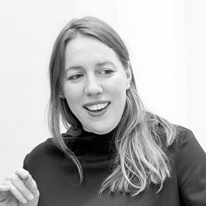 Black and white image of a person with long hair smiling and looking to the side. They are wearing a dark top and seem to be engaged in a conversation or activity. The background is plain and light-colored.