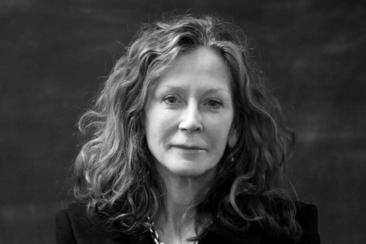 Black-and-white portrait of a middle-aged woman with wavy hair, looking directly at the camera with a neutral expression. She is wearing a dark top and a necklace, positioned against a plain, dark background.