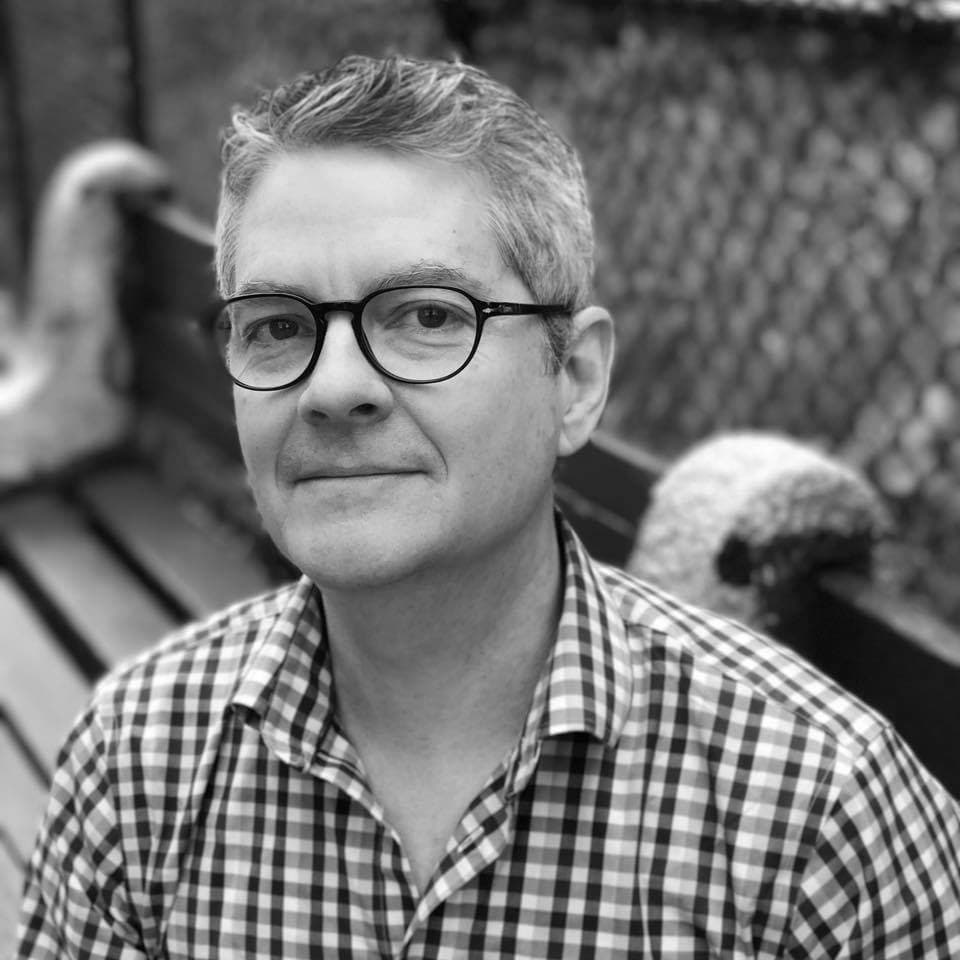 Black and white portrait of a man wearing glasses and a checkered shirt, sitting on a bench with a chain-link fence in the background.