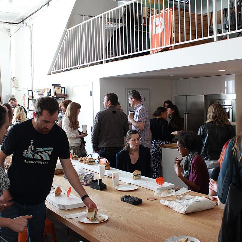 A group of people socialize and enjoy food in a modern, industrial-style space. Some are seated, while others stand near a long wooden table with snacks and beverages. A mezzanine level is visible above, and the atmosphere is casual and lively.