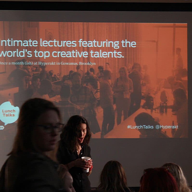 A presentation screen displays the text: "Intimate lectures featuring the world's top creative talents. Once a month(ish) at Hyperakt in Gowanus, Brooklyn." The room is dimly lit with several people gathered and conversing, some holding drinks.