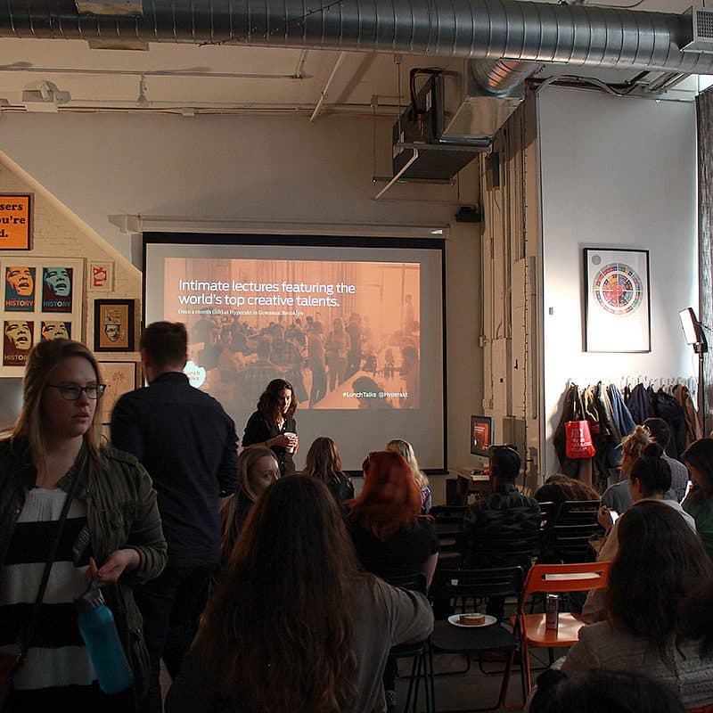 A group of people sitting in a dimly lit room watching a presentation projected on a screen. The slide reads "Intimate lectures featuring the world's top creative talents." Various posters adorn the walls, and the audience appears attentive.
