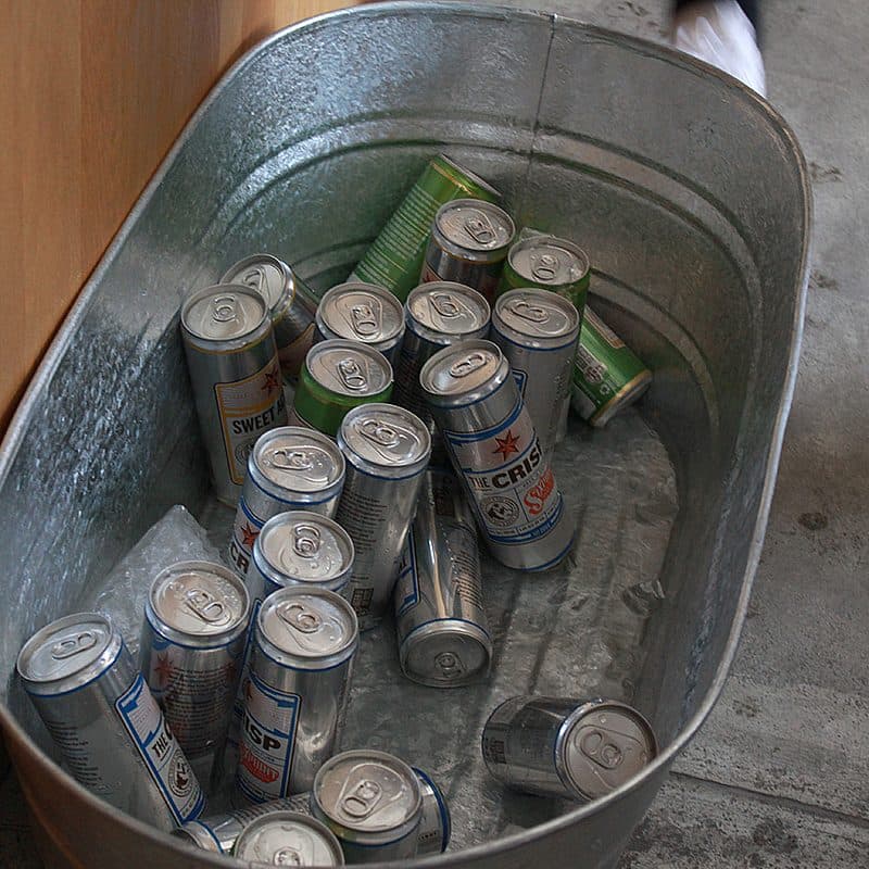 A large, oval-shaped metal tub filled with several empty beer cans of different brands is placed on a concrete floor. The tub is partially surrounded by a wooden surface, and a person's foot is visible near the top right corner of the image.