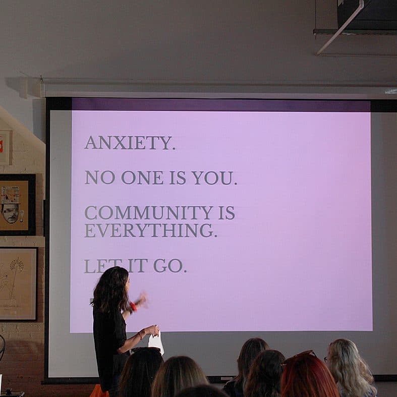 A presenter stands in front of a screen displaying text that reads, "Anxiety. No one is you. Community is everything. Let it go." There is an audience facing the screen, and the room has various artworks on the walls.