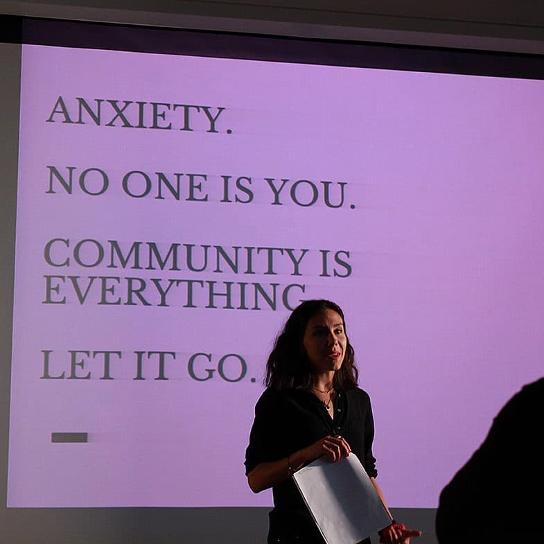 A woman stands in front of a projector screen giving a presentation. The screen displays text reading: "ANXIETY. NO ONE IS YOU. COMMUNITY IS EVERYTHING. LET IT GO." The background is dimly lit, and another person's silhouette is visible in the foreground.