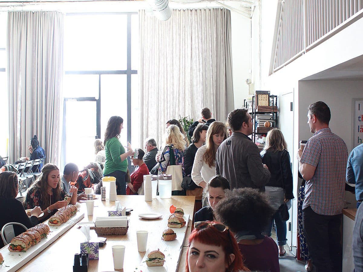 A group of people are gathered indoors in a bright, airy space with large windows. Some are seated around a long wooden table with food and drinks, while others stand and chat. The atmosphere appears social and casual, with a mix of relaxed and active interactions.