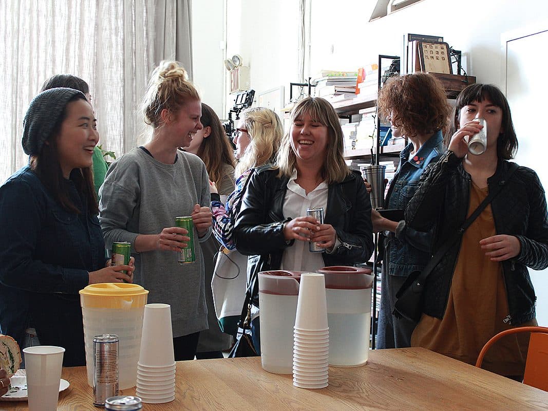 A group of people are gathered in a room, socializing and enjoying drinks. Some are holding cans while others sip from cups. There are pitchers and disposable cups on the table in the foreground. Shelves with books and other items are visible in the background.