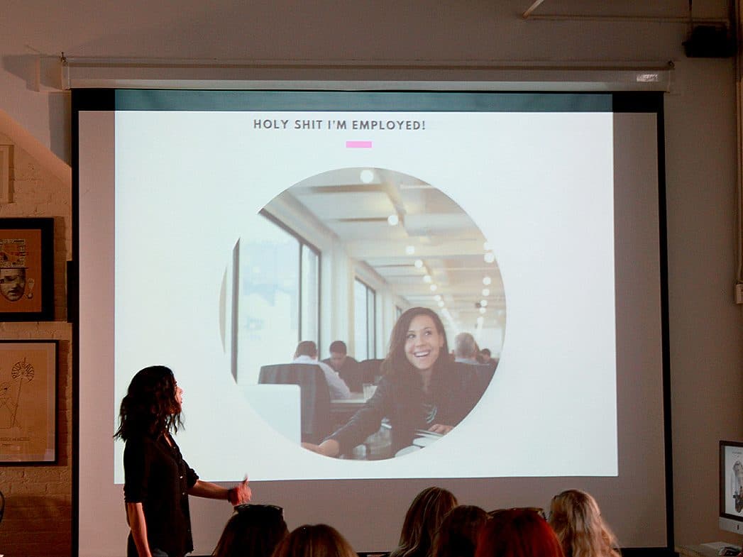 A person is giving a presentation in front of a group. On the screen behind them, there is a large image of another person smiling with the text "HOLY SHIT I'M EMPLOYED!" The audience is seated, and various artworks are visible on the walls.