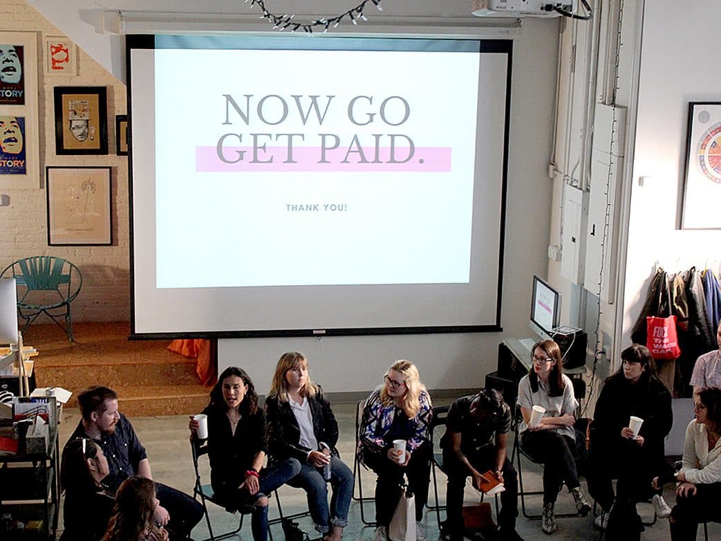 A group of people are sitting in chairs in a room, facing a large screen that displays the text "NOW GO GET PAID." The room has various posters and decorations on the walls. One person is speaking into a microphone while others listen and some drink from cups.