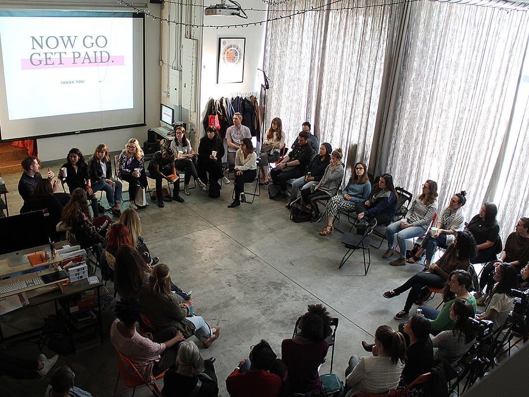 A large group of people are seated in a circle in a spacious room with high ceilings. A projector screen reads "NOW GO GET PAID." The attendees appear to be engaged in a discussion or workshop. Light streams in from tall windows on the right side of the room.