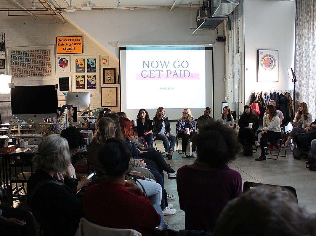 A group of people are sitting in a spacious, well-lit room arranged in a circle. Above the participants, a projector screen displays the words "NOW GO GET PAID." The space has eclectic décor, including framed posters and various artworks on the walls.