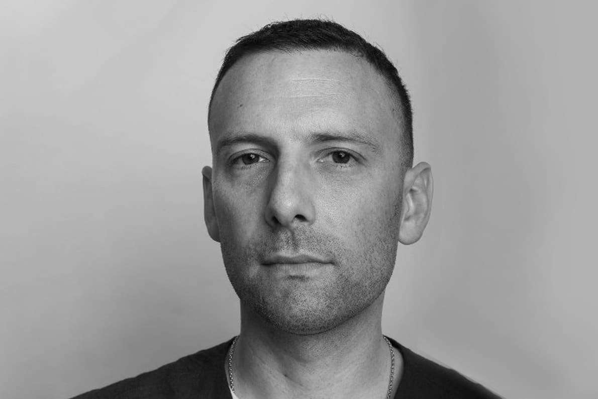 A black and white portrait of a man with short hair, wearing a simple shirt. He has a slight facial hair and neutral expression while looking directly at the camera. The background is plain and gradient from light to dark.