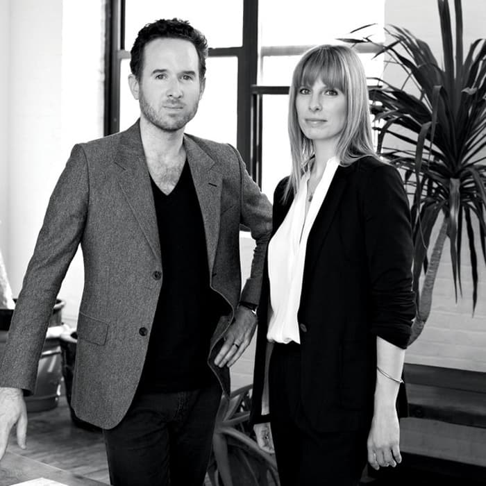 A black and white photo of a man and woman standing indoors beside a large window. The man on the left is wearing a blazer over a V-neck shirt, while the woman on the right is dressed in a blazer over a blouse. There is a plant in the background.