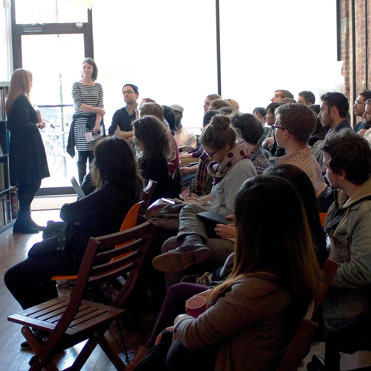 A room filled with adults seated on chairs, attentively listening to two standing presenters. The room is lit by natural light streaming in from large windows. The setup suggests an informal, interactive seminar or workshop setting, with a relaxed and engaged audience.
