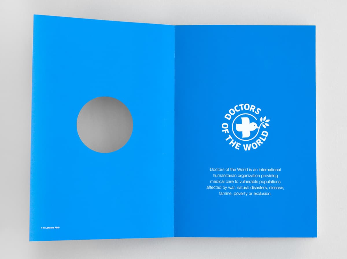 Open book with a blue page on the right side. The right page features the white logo of "Doctors of the World," a circle with a cross and dove, and text describing the organization as providing medical care to vulnerable populations affected by various hardships.