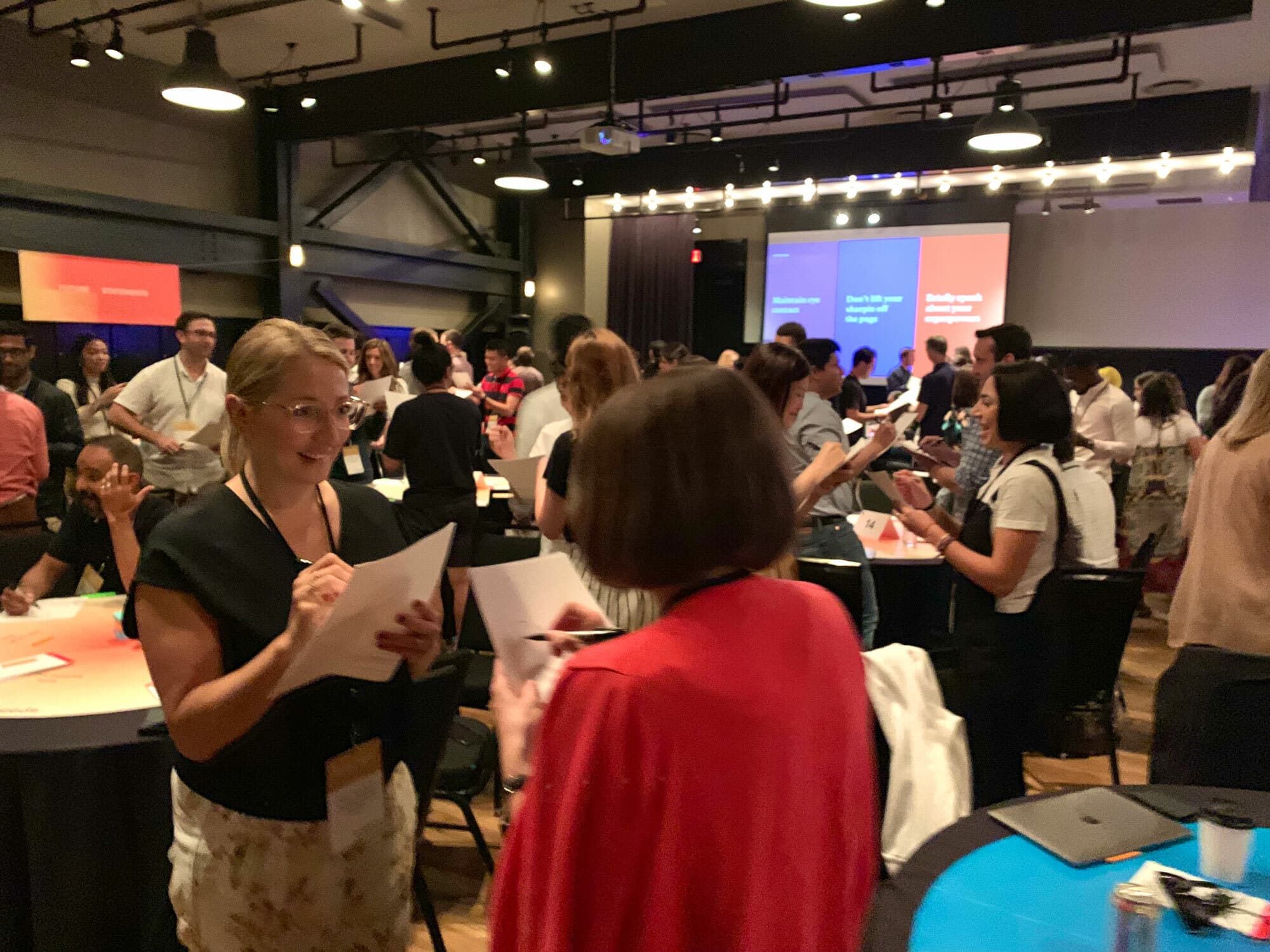 People are gathered in a large room engaging in discussions and writing on papers. Some are standing near tables, while others are mingling. The room has modern lighting, a stage with screens displaying text, and a lively, collaborative atmosphere.