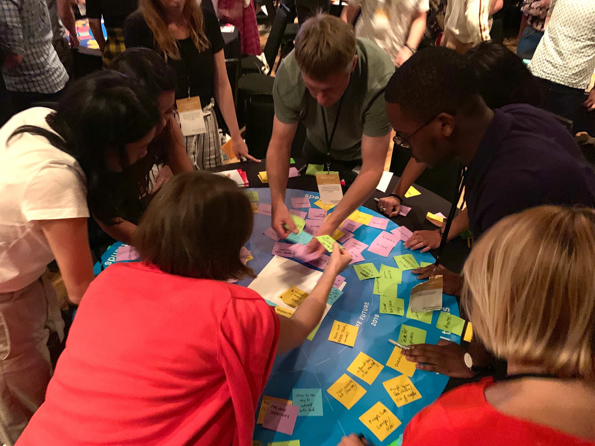 A group of people gathered around a table participating in a collaborative activity with colorful sticky notes. The scene appears dynamic, with each person engaged in adding or rearranging notes on a large sheet.