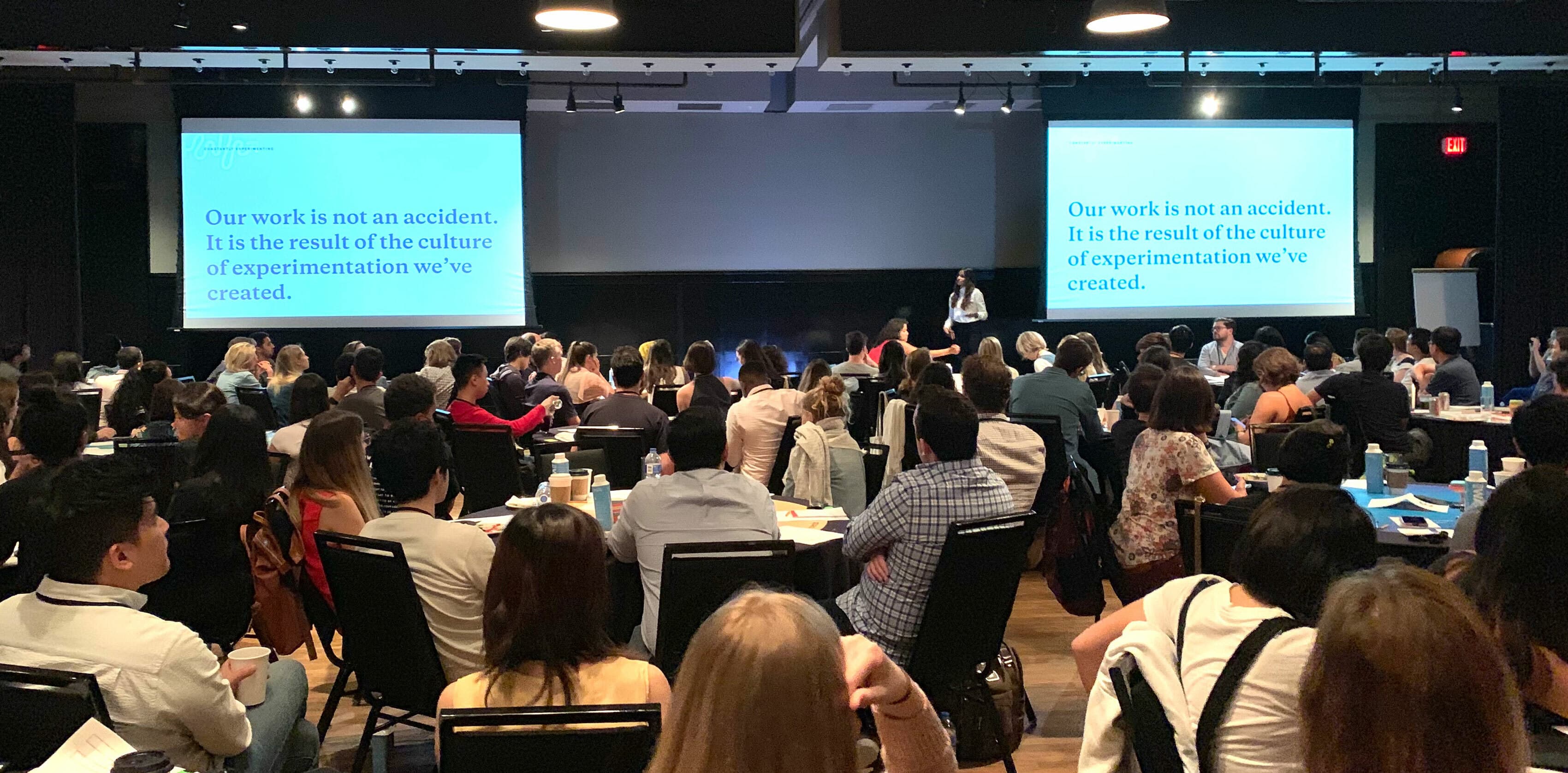 A speaker stands in front of a large audience in a conference room, presenting with the aid of two large screens displaying the text: "Our work is not an accident. It is the result of the culture of experimentation we've created." The audience is seated and attentive.