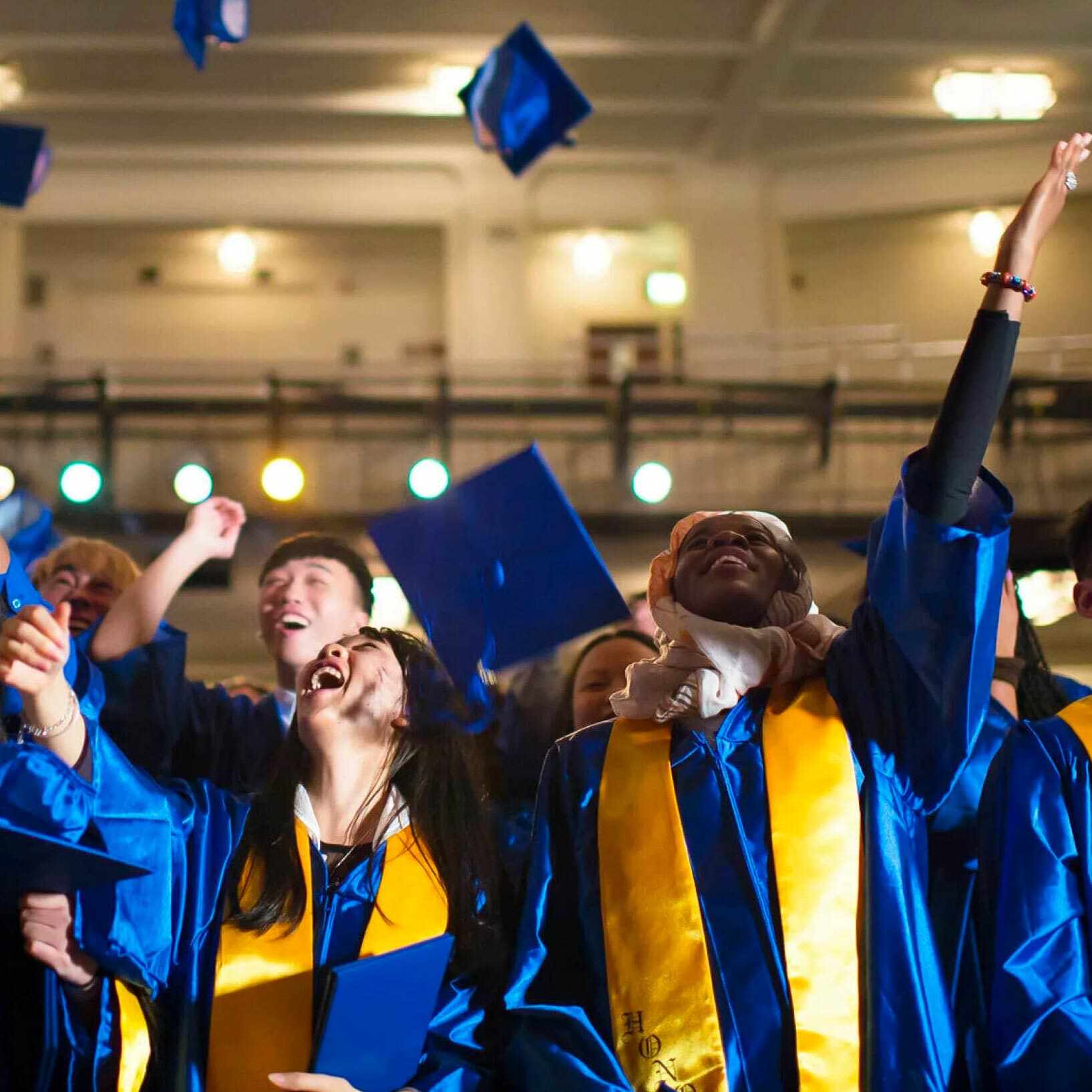 Graduates in blue gowns and caps celebrate by throwing their caps in the air. They smile and raise their hands joyfully. The background features an indoor venue with lights and seating areas.