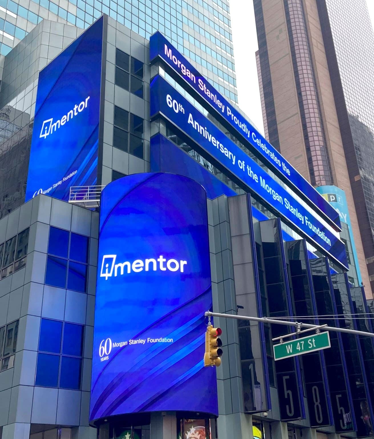 Digital billboards on a tall building display messages celebrating the 60th anniversary of the Morgan Stanley Foundation. The words "4 mentor" and "Morgan Stanley Proudly Celebrates the 60th Anniversary of the Morgan Stanley Foundation" are prominently visible.