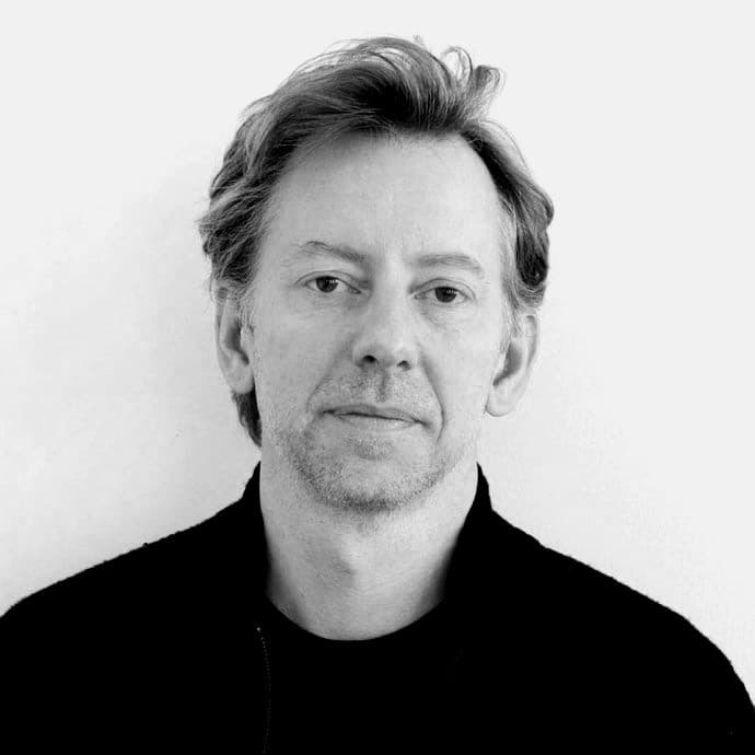 A black-and-white portrait of a man with medium-length hair, wearing a dark jacket over a black shirt. He has a neutral expression and is looking directly at the camera. The background is plain white.