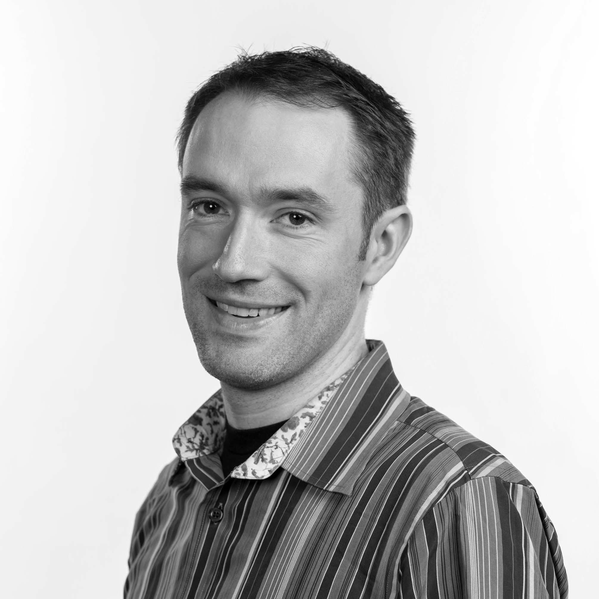 A black and white photo of a smiling person with short, dark hair. They are wearing a striped shirt with a light-colored floral collar. The background is plain white.