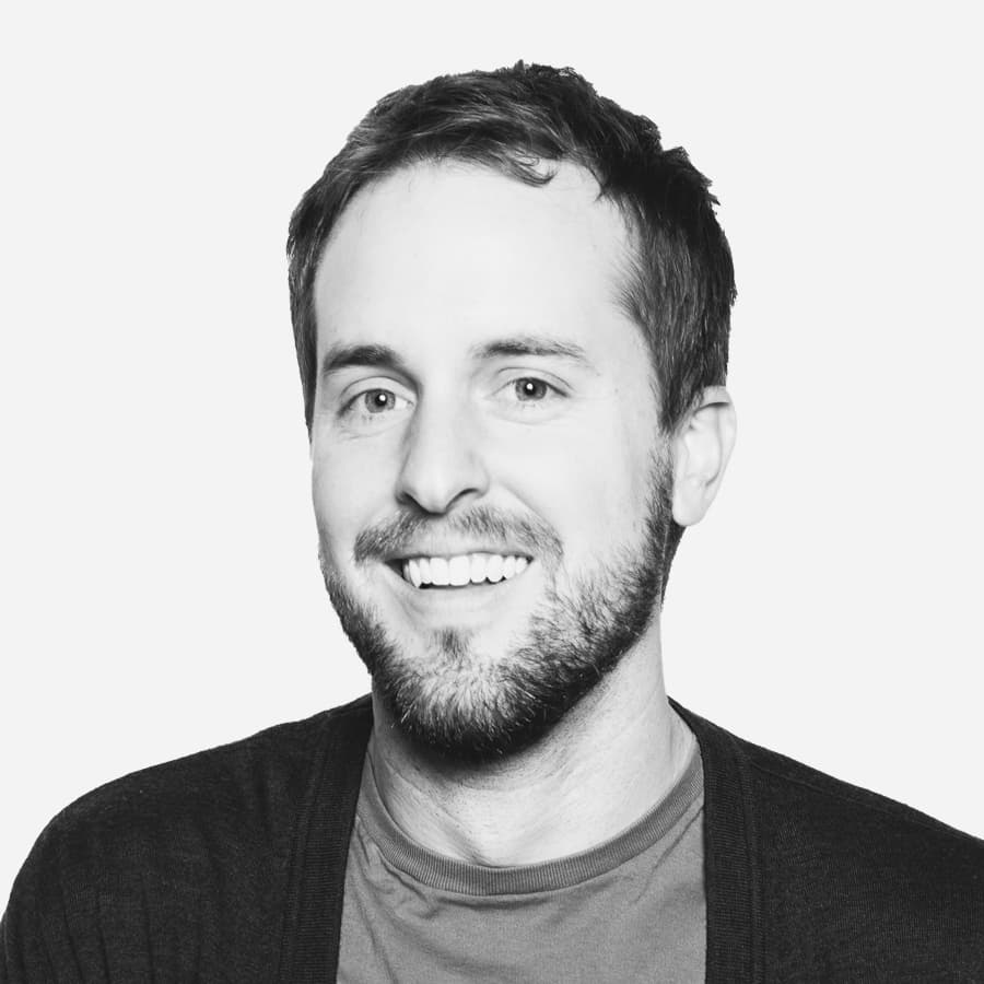 A man with short, dark hair and a beard is smiling. He is wearing a dark cardigan over a t-shirt. The background is plain and light-colored.