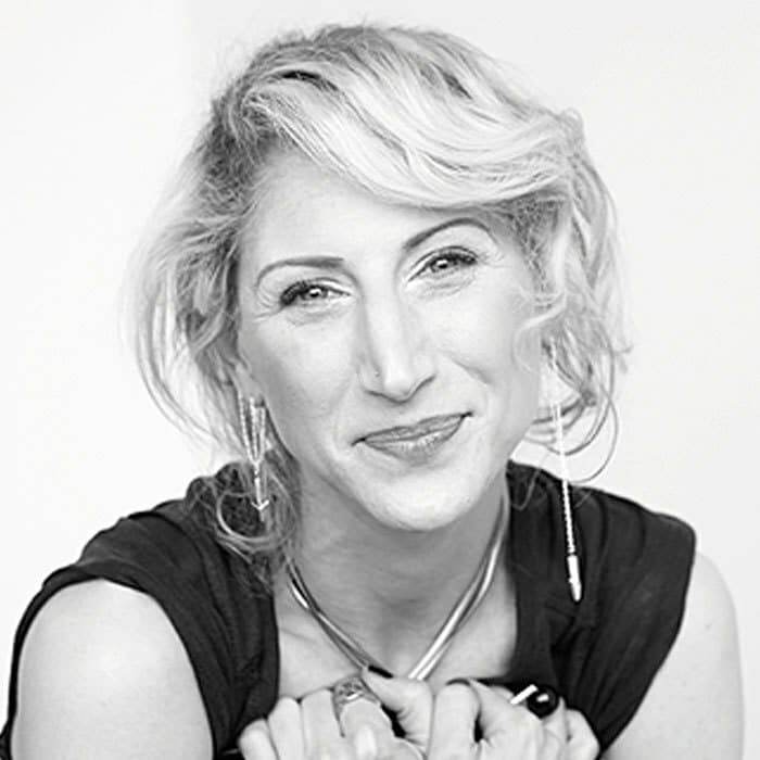 Black and white photo of a smiling person with short, wavy, light-colored hair. They are wearing a sleeveless top, long earrings, and a thick necklace. Their hands are clasped together, resting under their chin. The background is plain and light-colored.