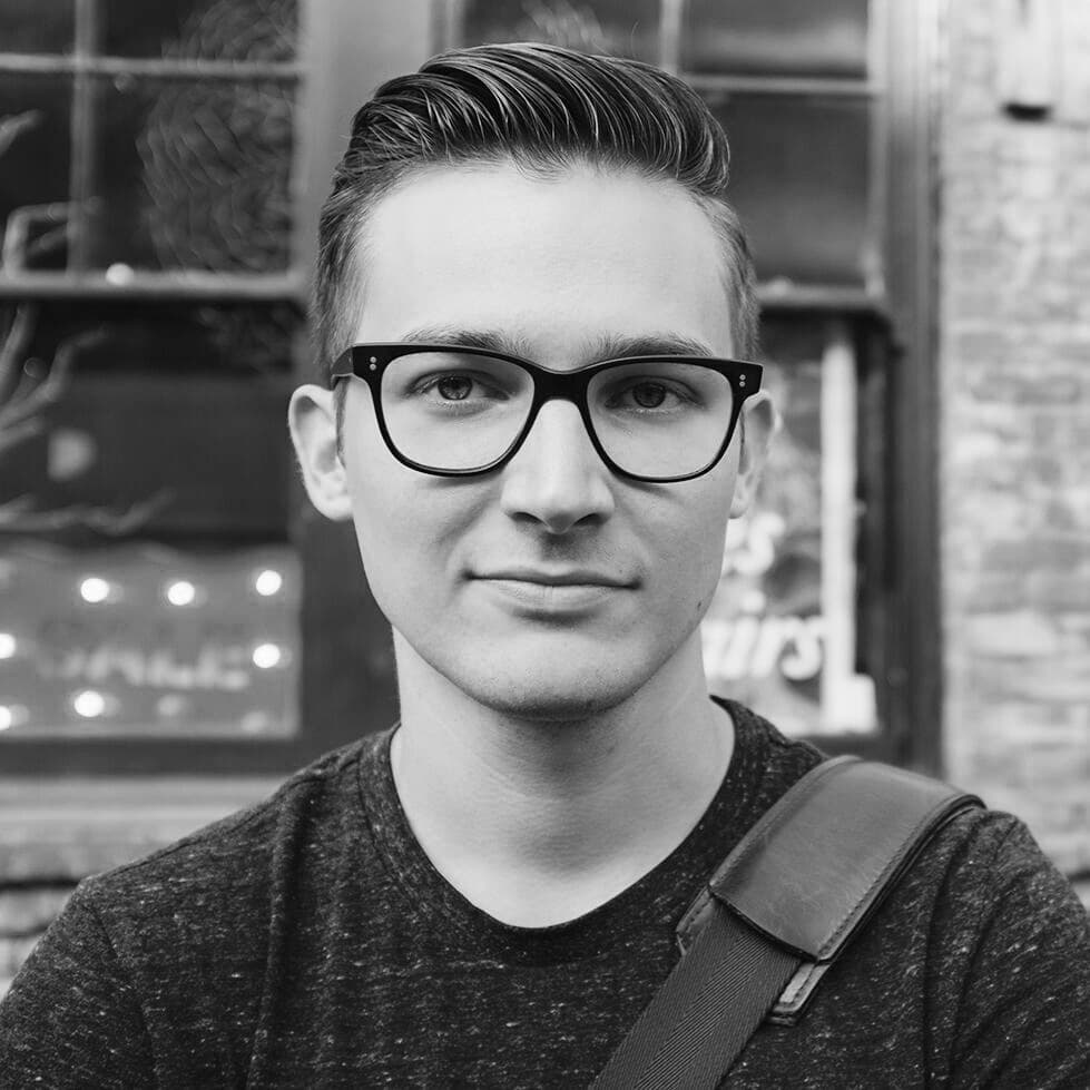 Black and white photo of a person with short, neatly styled hair and wearing black-rimmed glasses. They are dressed in a plain dark t-shirt with a strap over their shoulder, standing in front of a building with a window displaying faint signage.