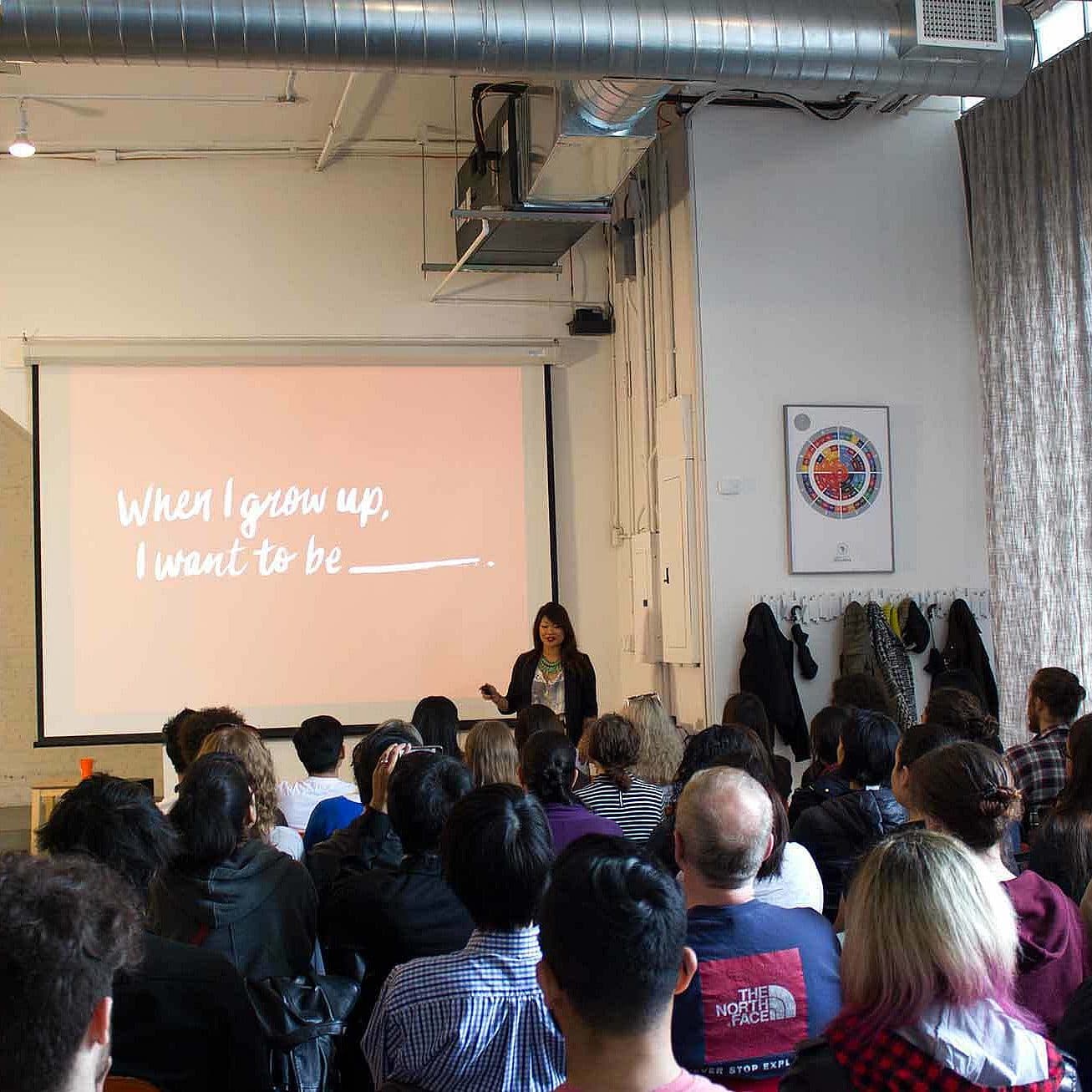 A woman is giving a presentation to a seated audience in a modern room. Behind her, a large screen displays the text, "When I grow up, I want to be _______." The attendees are attentively facing the speaker. The room is illuminated with natural light.