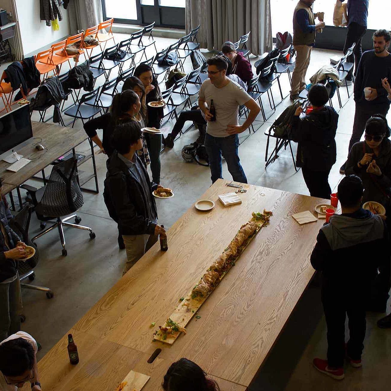 A group of people are gathered around a long wooden table covered with food at a casual indoor event. Some attendees are standing and holding plates with food, while others are sitting or standing nearby. Several chairs are scattered around the room.