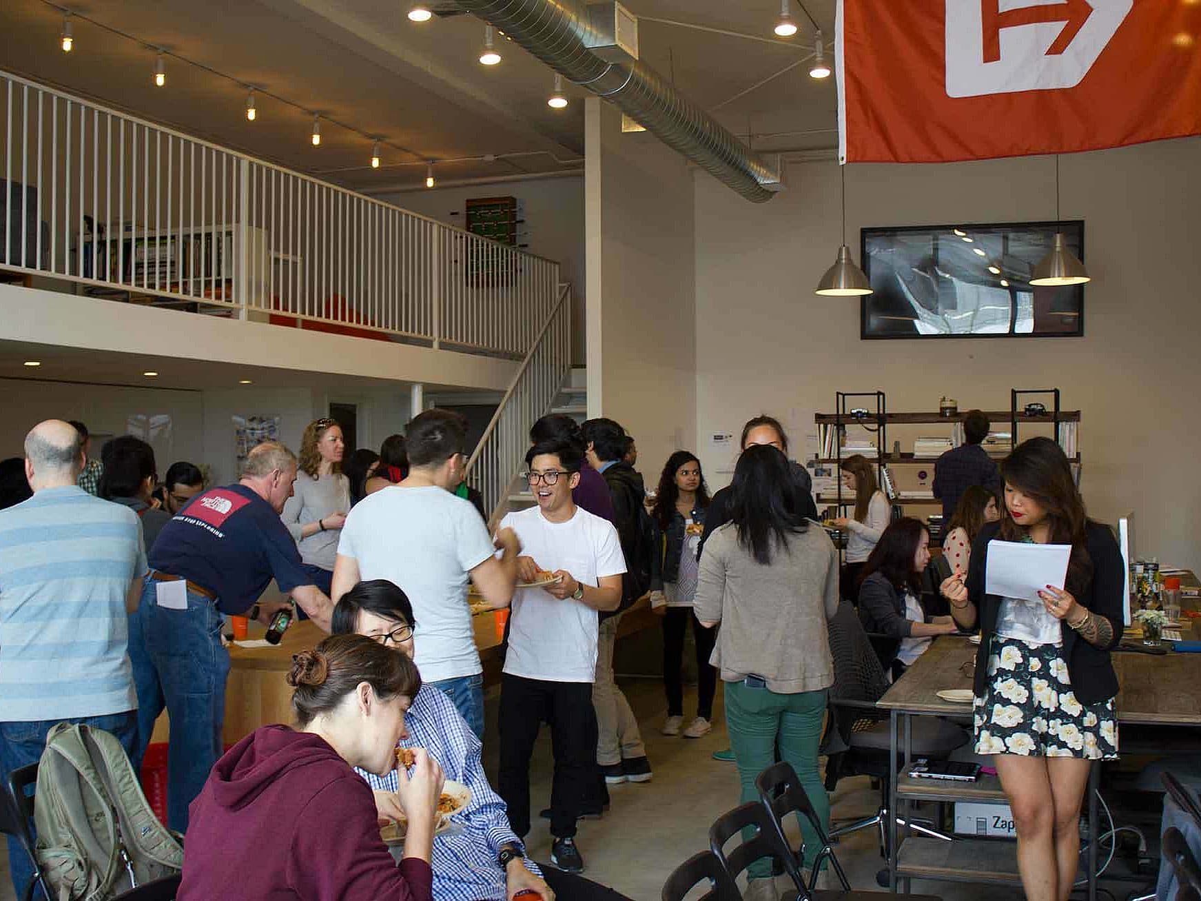 A diverse group of people socializing and enjoying a gathering in a modern, open-layout space with high ceilings and string lighting. Some individuals are standing and chatting, while others are seated at tables. A loft area and a hanging orange banner are visible in the background.
