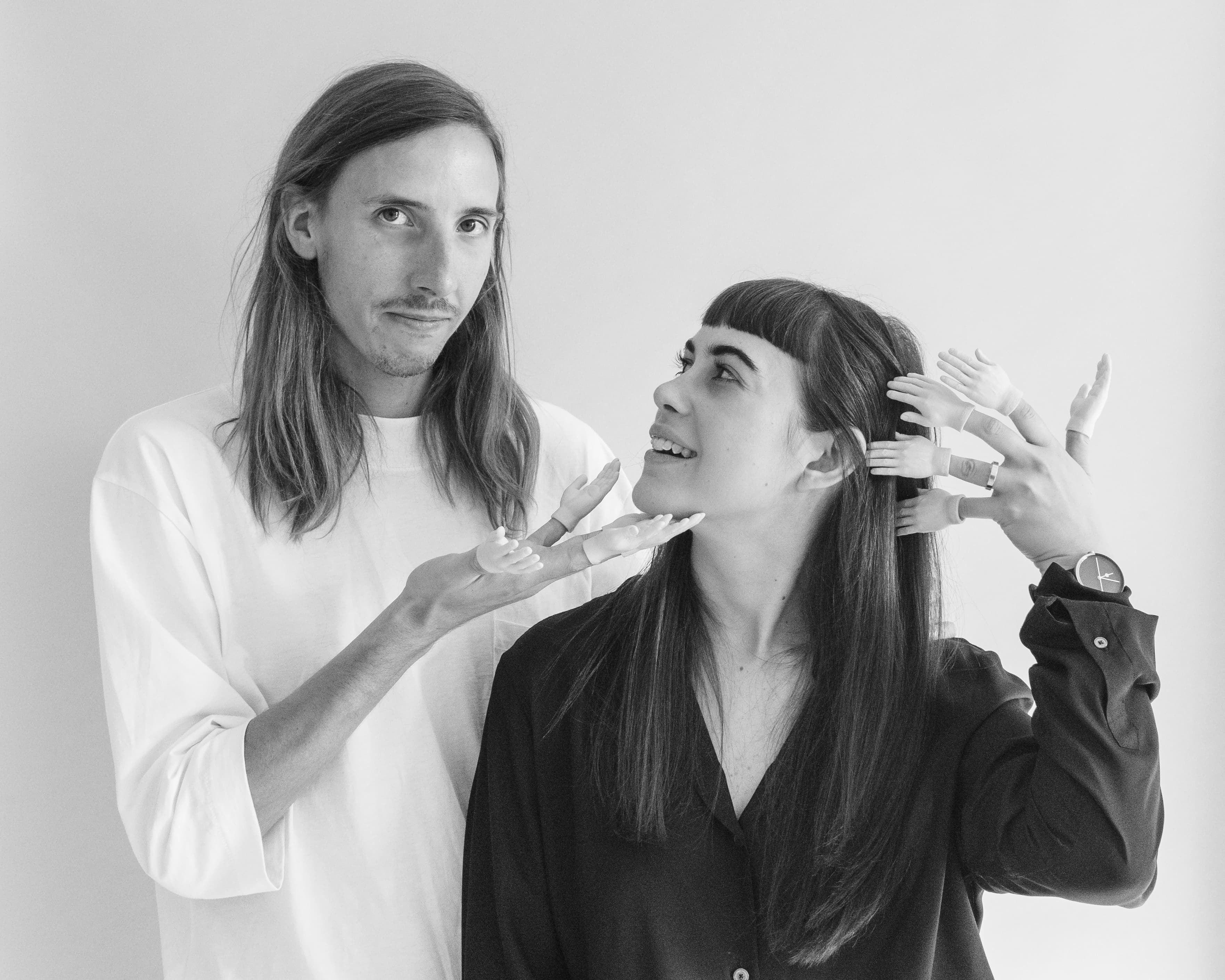 A man with long hair and a woman with bangs, both dressed in casual clothes, stand against a plain background. The woman has small toy hands on her fingers, while the man playfully gestures toward her. Both are smiling and looking at each other. Black and white photo.