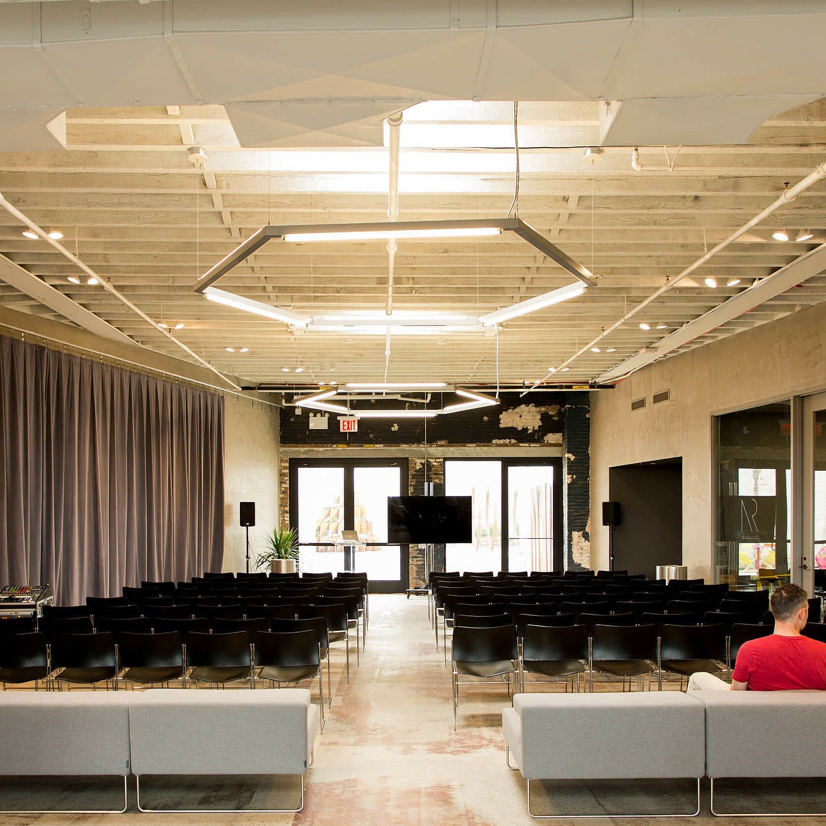 A modern conference room with rows of black chairs facing a large television screen at the front. Two people sit on gray sofas at the back of the room. The room has a high ceiling, exposed ductwork, and large windows allowing natural light.