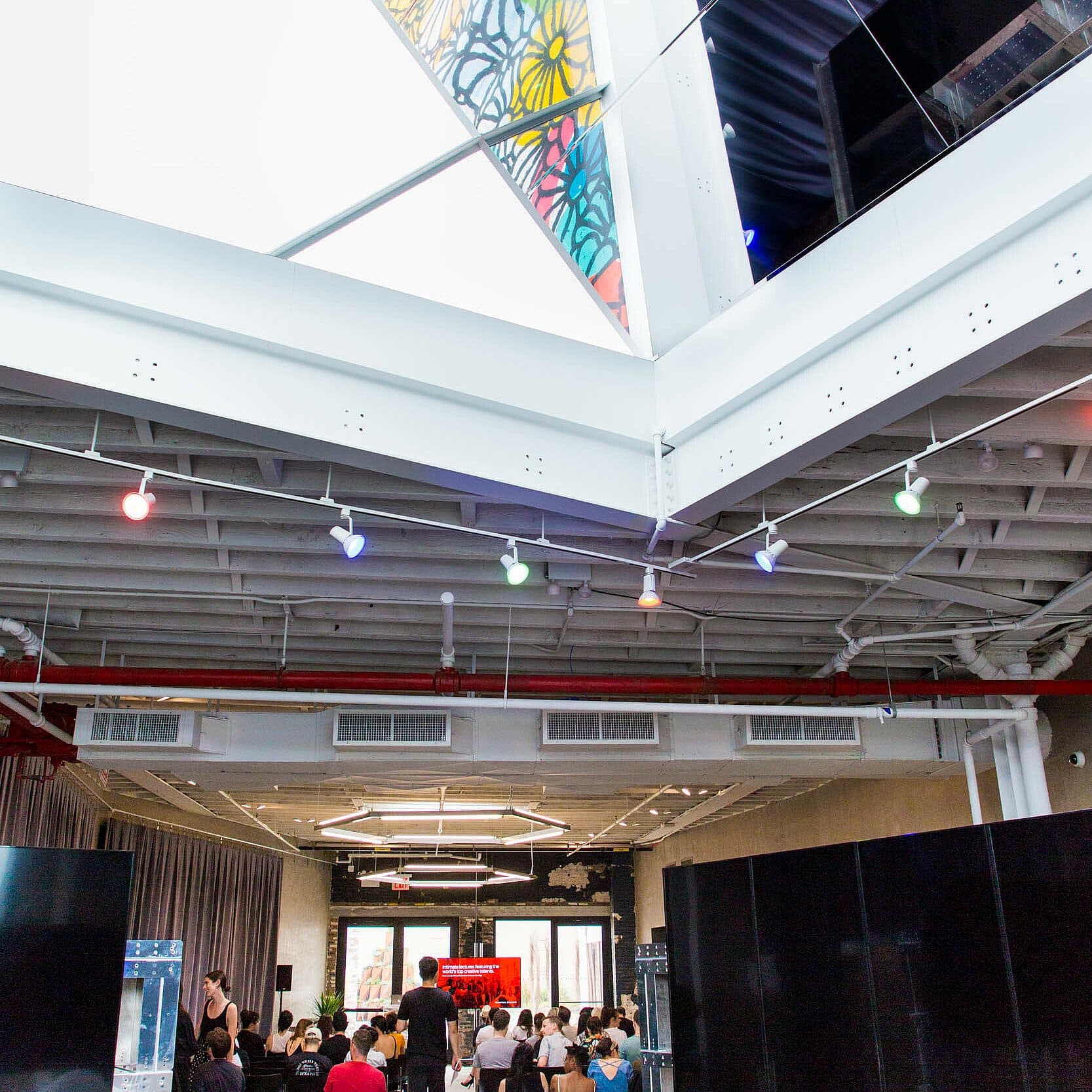 A modern indoor space with a high ceiling and large skylights adorned with colorful, floral-themed stained glass. Below, a group of people is seated and facing a raised platform, with several screens and spotlights around the room.