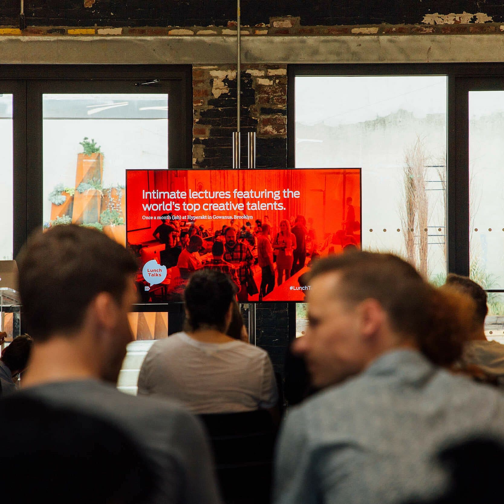 A group of people is seated in a room, facing a presentation screen. The screen displays text that reads, "Intimate lectures featuring the world's top creative talents." The setting appears informal, with a mix of natural and indoor light.
