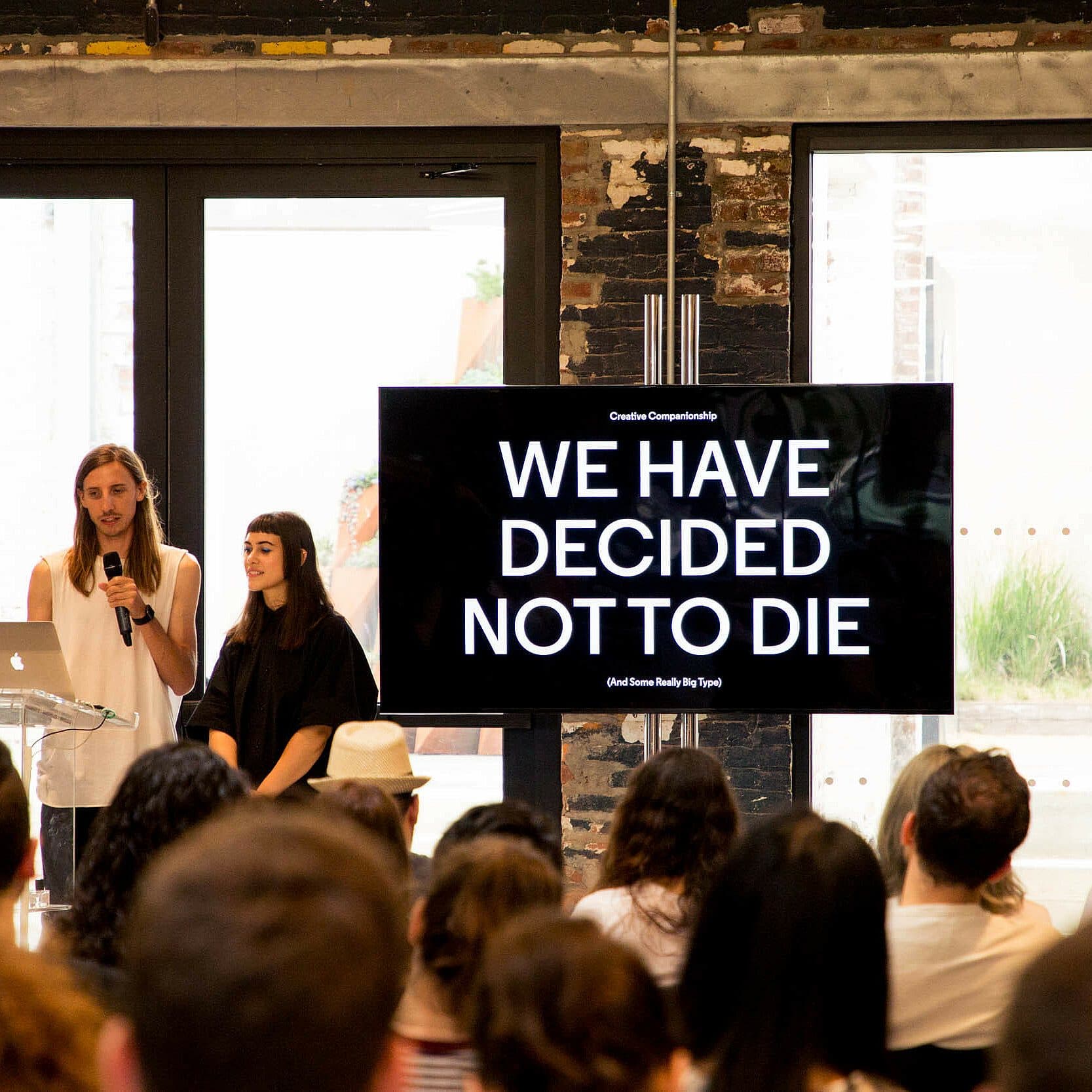 Two individuals stand in front of a seated audience, presenting with a microphone and screen. The screen displays the text "WE HAVE DECIDED NOT TO DIE". They are in a room with large windows and a brick wall backdrop.
