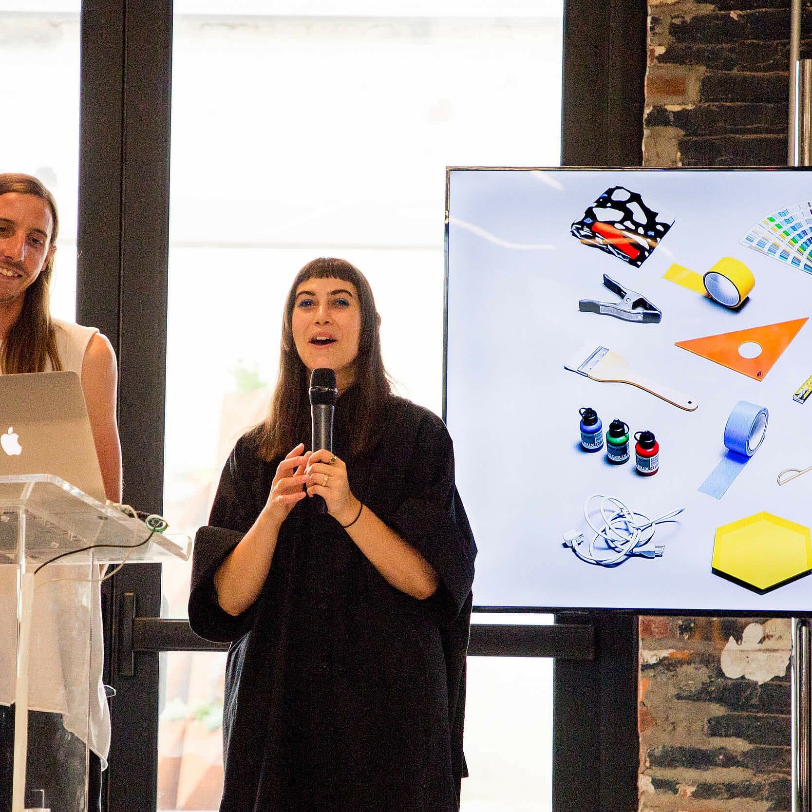Two people are presenting in front of an audience. The person on the left stands behind a clear podium with a laptop. The person on the right holds a microphone. A large screen displays various colorful stationery items and tools. The background has large windows.