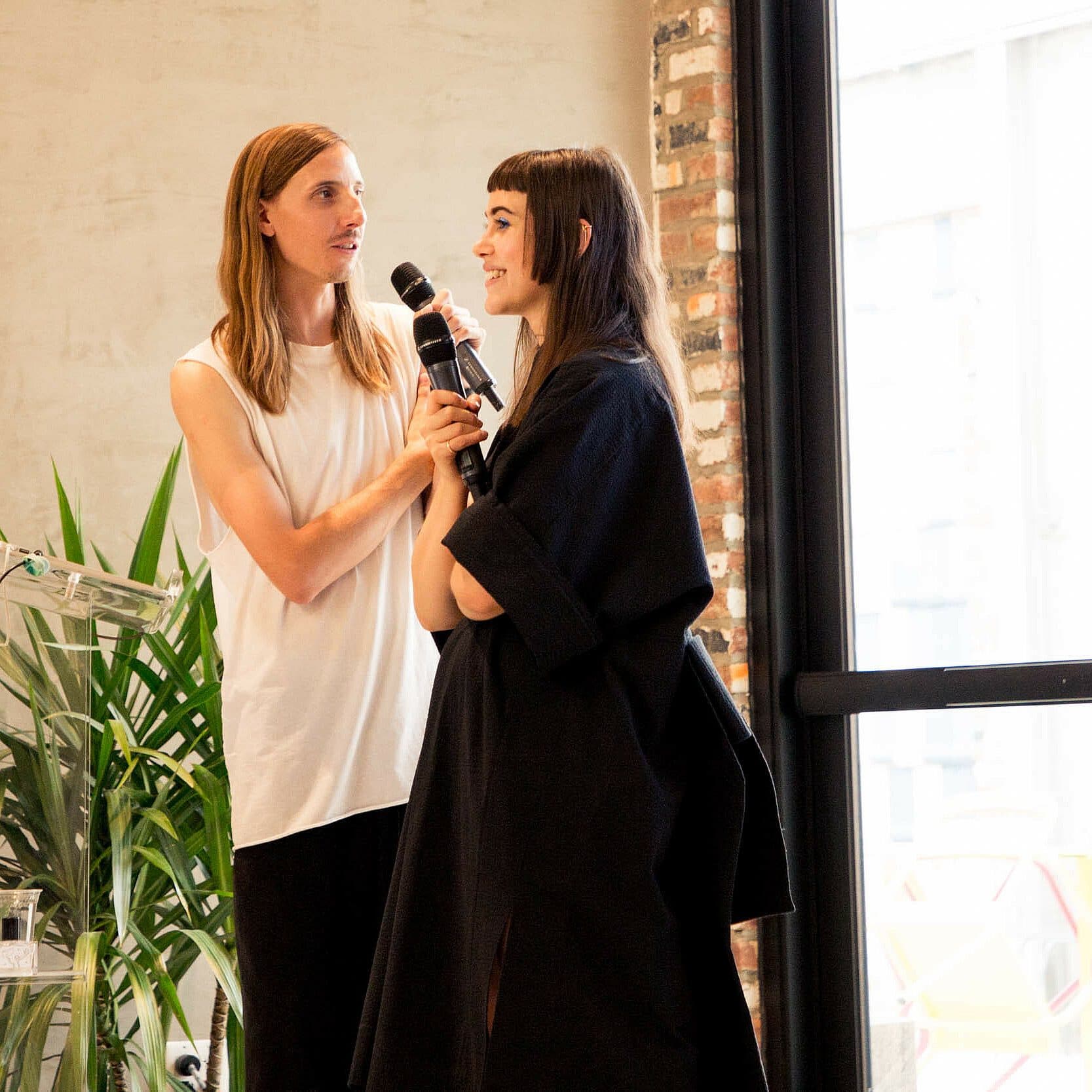 Two people stand indoors by a window, engaged in conversation. The person on the left has long hair, is wearing a white sleeveless top, and is holding a microphone. The person on the right has dark hair, is wearing a dark outfit, and is smiling. A plant stands nearby.